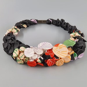 Puede incluir: Un collar de tela negro con un grupo de flores de tela de colores. Las flores están hechas de diferentes telas con varios patrones, incluyendo rayas, lunares e estampados florales. El collar es una pieza llamativa con un diseño atrevido y único.