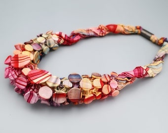 Puede incluir: Un collar colorido con base de tela y adornos circulares estampados. El collar presenta una variedad de colores, incluyendo rojo, rosa, amarillo y azul. El cierre es de metal oscuro. El diseño es artístico y llamativo.