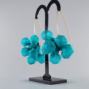 Peut inclure: Boucles d'oreilles en tissu bleu sarcelle avec des fils d'oreille dorés. Les boucles d'oreilles sont présentées sur un présentoir noir sur fond gris. Les boucles d'oreilles ont un design artistique unique.