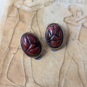 May include: A pair of oval-shaped clip-on earrings featuring dark red, carved scarab beetle motifs set in polished gold-tone metal frames. The scarab designs display detailed horizontal and vertical lines, creating a distinct insect-like appearance.
