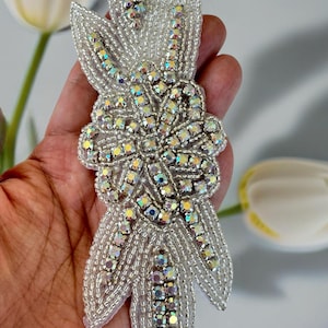 May include: A silver beaded and rhinestone applique with a floral design. The applique is made of small, iridescent rhinestones and silver beads.