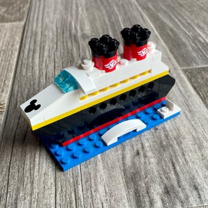 May include: A white, black, and red Lego model of a cruise ship with Mickey Mouse ears on the front. The ship has two red funnels with black tops and a white deck with yellow accents. The ship is sitting on a blue Lego base.
