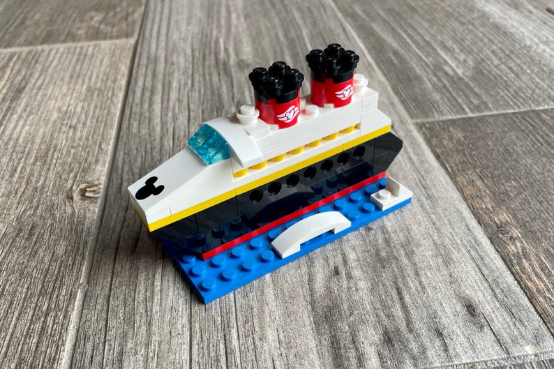 May include: A white, black, and red Lego model of a cruise ship with Mickey Mouse ears on the front. The ship has two red funnels with black tops and a white deck with yellow accents. The ship is sitting on a blue Lego base.