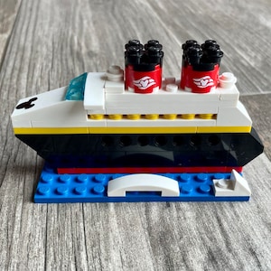May include: A white, black, and red Lego model of a cruise ship with Mickey Mouse ears on the bow. The ship has two red funnels with white Mickey Mouse heads and a yellow stripe along the side. The ship is sitting on a blue Lego base.