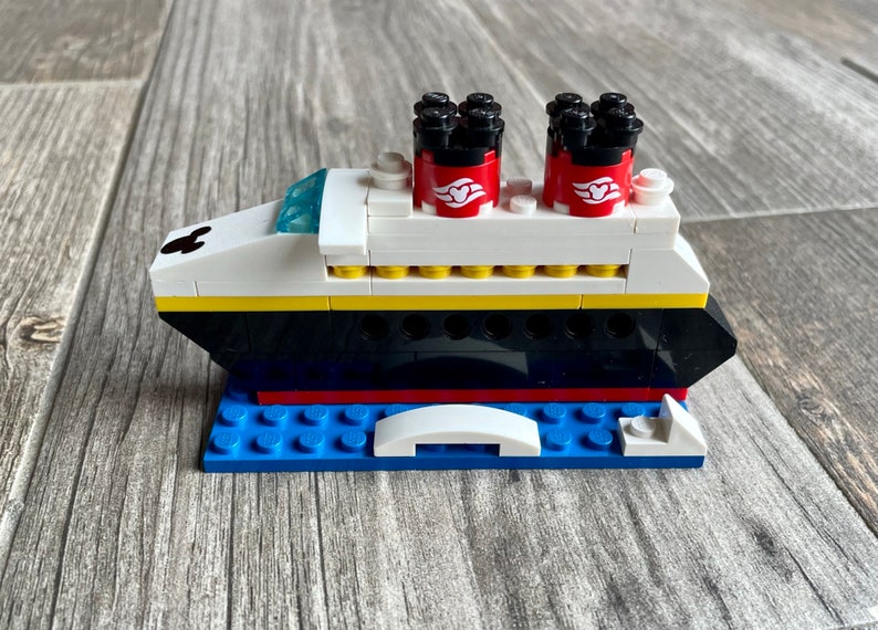 May include: A white, black, and red Lego model of a cruise ship with Mickey Mouse ears on the bow. The ship has two red funnels with white Mickey Mouse heads and a yellow stripe along the side. The ship is sitting on a blue Lego base.