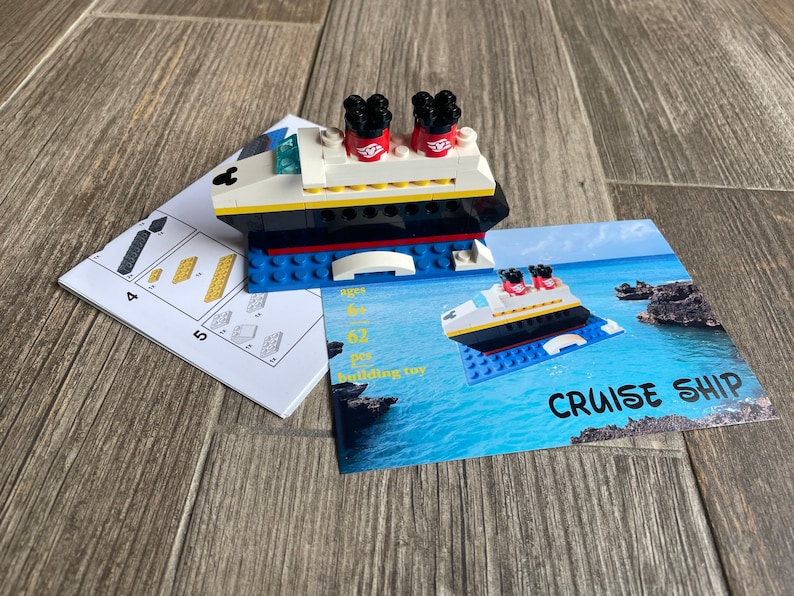 May include: A small, blue, black, and white LEGO cruise ship building toy with red funnels. The ship is on a blue base with a white Mickey Mouse head on the side. The toy is next to a set of instructions and a picture of the finished ship.