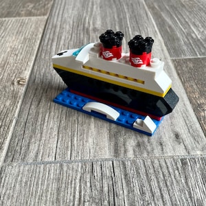 May include: A small, white and black Lego model of a cruise ship with red and black smokestacks. The ship is sitting on a blue Lego base.