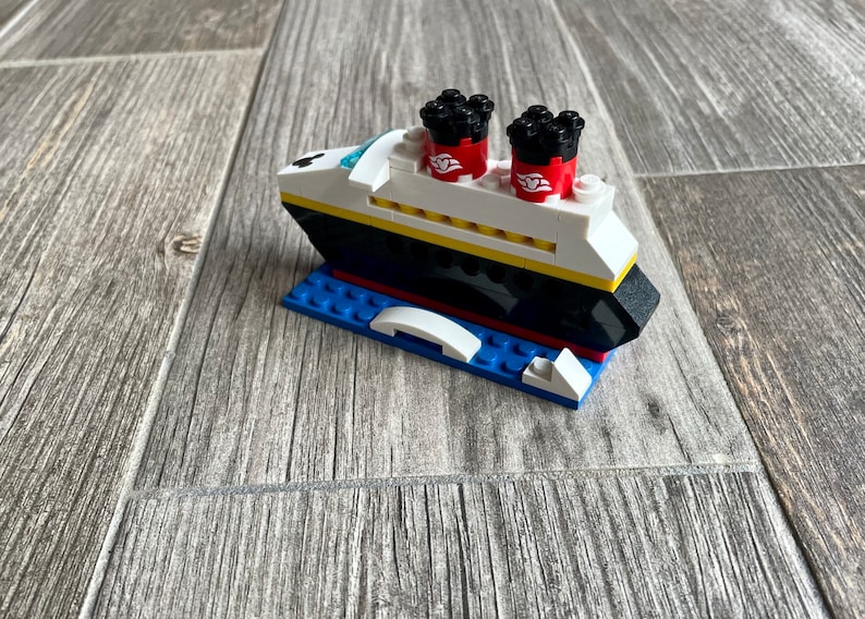May include: A small, white and black Lego model of a cruise ship with red and black smokestacks. The ship is sitting on a blue Lego base.