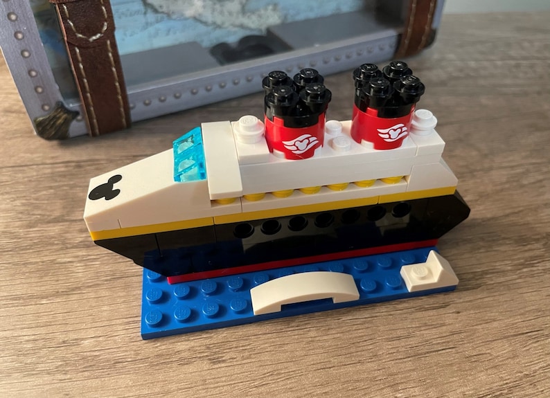 May include: A white, black, and yellow LEGO model of a cruise ship with red funnels and a Mickey Mouse head on the front. The ship is sitting on a blue LEGO base.