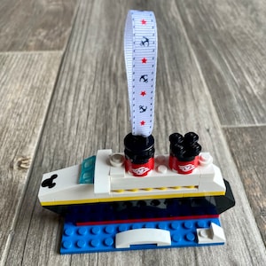 May include: A white, black, and yellow Lego ornament of a cruise ship with red Mickey Mouse ears and a white ribbon with red stars and anchors.