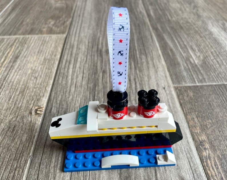 May include: A white, black, and yellow Lego ornament of a cruise ship with red Mickey Mouse ears and a white ribbon with red stars and anchors.