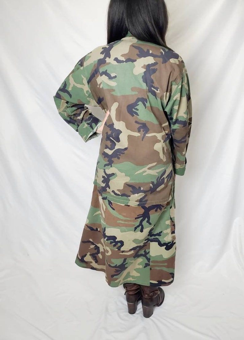 Pode incluir: Casaco comprido com padr&atilde;o de camuflagem em tons de verde, castanho e preto. O casaco tem mangas compridas e uma parte inferior semelhante a uma saia. A pessoa est&aacute; a usar botas castanhas de salto alto. &Eacute; uma pe&ccedil;a de inspira&ccedil;&atilde;o militar.