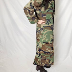 Pode incluir: Um casaco longo com padr&atilde;o de camuflagem com v&aacute;rios bolsos. O casaco tem uma frente abotoada e um decote com gola. As cores incluem tons de verde, castanho e preto. A pessoa est&aacute; a usar botas castanhas de salto alto.