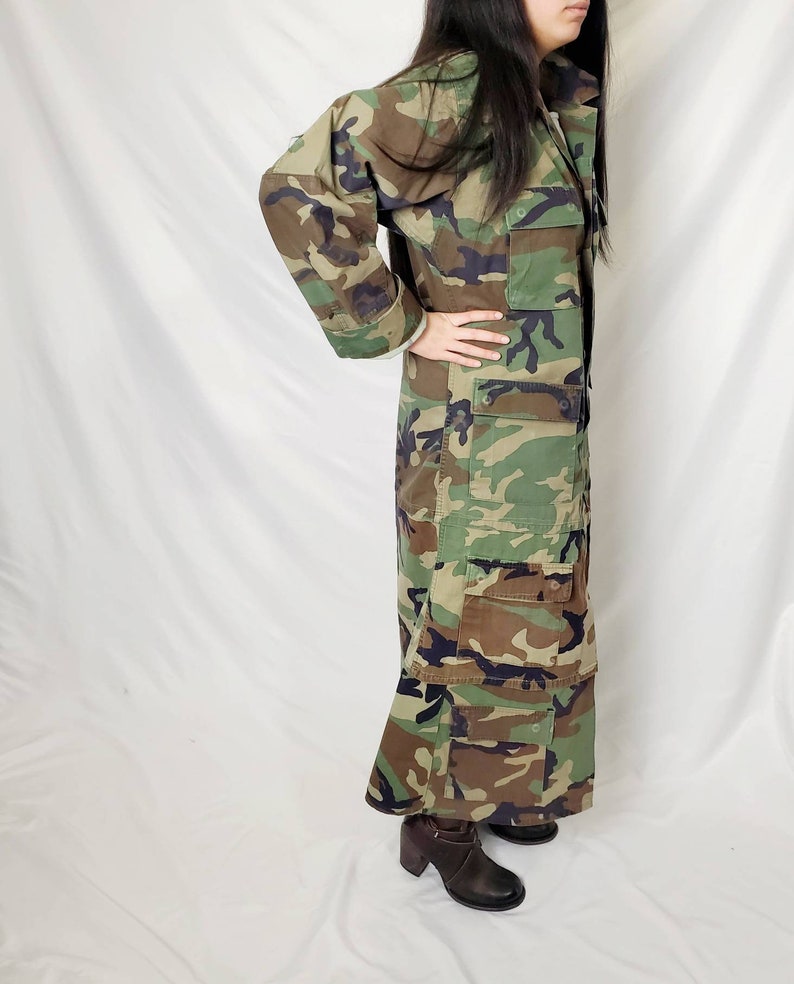 Pode incluir: Um casaco longo com padr&atilde;o de camuflagem com v&aacute;rios bolsos. O casaco tem uma frente abotoada e um decote com gola. As cores incluem tons de verde, castanho e preto. A pessoa est&aacute; a usar botas castanhas de salto alto.