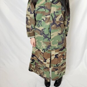 Pode incluir: Um casaco longo de camuflagem em tons de verde, castanho e preto. O casaco apresenta v&aacute;rios bolsos e uma frente abotoada. A pe&ccedil;a de vestu&aacute;rio &eacute; at&eacute; ao joelho e tem um design de inspira&ccedil;&atilde;o militar.