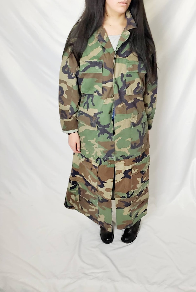 Pode incluir: Um casaco longo de camuflagem em tons de verde, castanho e preto. O casaco apresenta v&aacute;rios bolsos e uma frente abotoada. A pe&ccedil;a de vestu&aacute;rio &eacute; at&eacute; ao joelho e tem um design de inspira&ccedil;&atilde;o militar.
