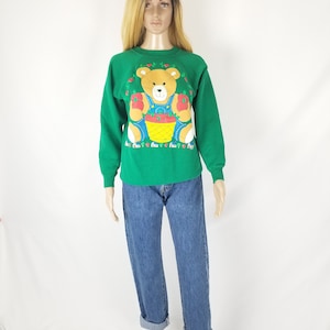 May include: Green sweatshirt with a cartoon teddy bear wearing overalls and holding a basket of strawberries. The bear is surrounded by small red strawberries and green leaves.