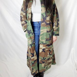 Pode incluir: Um casaco longo com padr&atilde;o de camuflagem em tons de verde, castanho e preto. O casaco tem v&aacute;rios bolsos e uma frente abotoada. A pessoa est&aacute; vestindo uma camisa cinza e jeans azuis. Sapatos pretos completam o look.