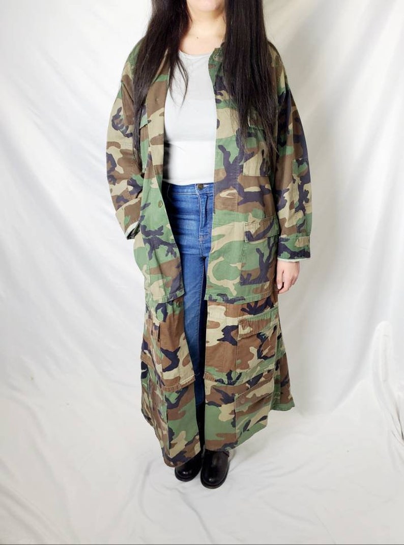 Pode incluir: Um casaco longo com padr&atilde;o de camuflagem em tons de verde, castanho e preto. O casaco tem v&aacute;rios bolsos e uma frente abotoada. A pessoa est&aacute; vestindo uma camisa cinza e jeans azuis. Sapatos pretos completam o look.