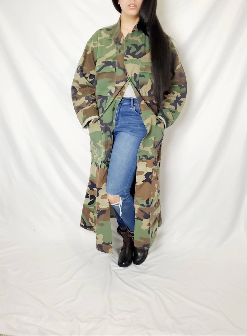 Pode incluir: Um casaco comprido com padr&atilde;o de camuflagem com v&aacute;rios bolsos. O casaco est&aacute; aberto, revelando um top cinzento e cal&ccedil;as de ganga azuis com um joelho rasgado. Botas pretas completam o look. As cores do casaco incluem tons de verde, castanho e preto.