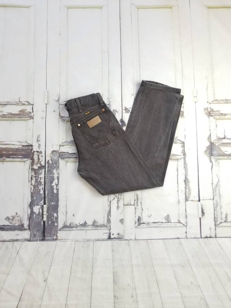 May include: A pair of dark gray denim jeans with a brown leather patch on the back pocket. The jeans are folded in half and laid flat on a white wooden surface.