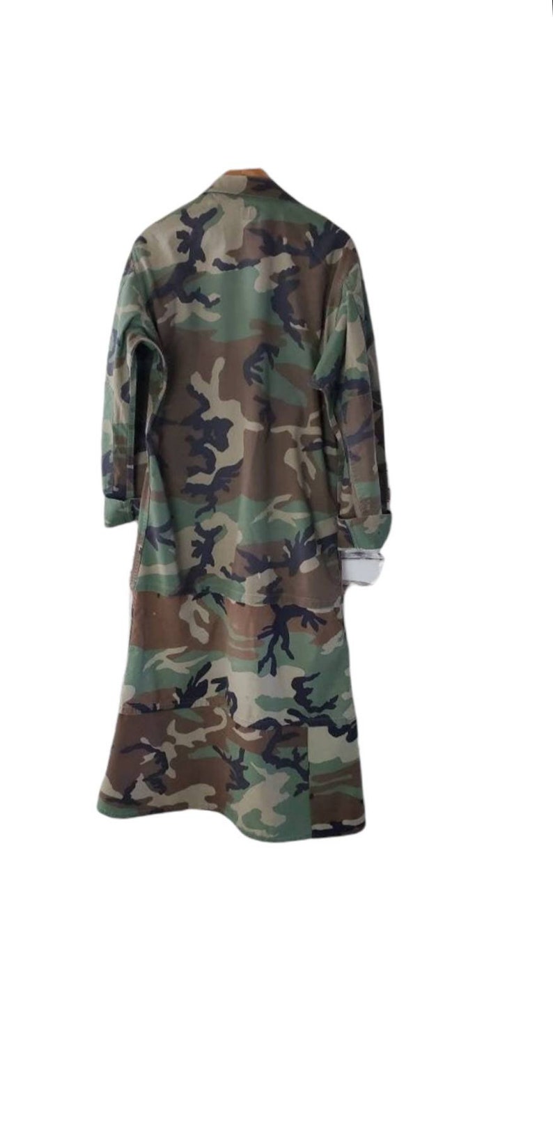 Pode incluir: Um casaco longo com padr&atilde;o de camuflagem. O casaco apresenta uma frente abotoada, mangas compridas e um colarinho. O design de camuflagem incorpora tons de verde, castanho e preto.
