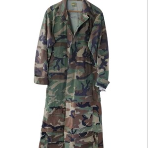 Pode incluir: Um casaco longo de camuflagem com fecho de bot&otilde;es, v&aacute;rios bolsos e gola. O casaco &eacute; feito de um padr&atilde;o de camuflagem verde, castanho e preto e est&aacute; pendurado num cabide de madeira.