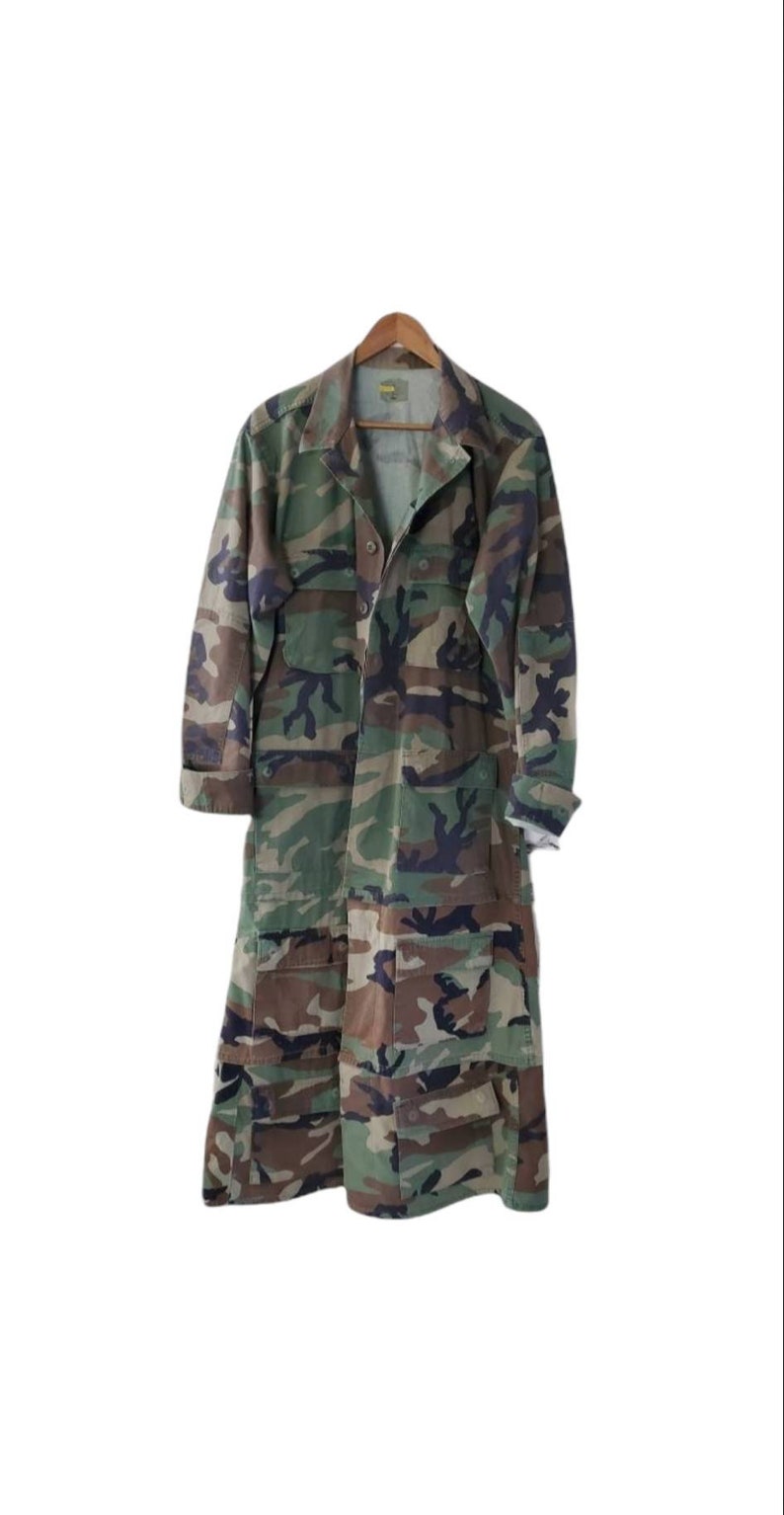 Pode incluir: Um casaco longo de camuflagem com fecho de bot&otilde;es, v&aacute;rios bolsos e gola. O casaco &eacute; feito de um padr&atilde;o de camuflagem verde, castanho e preto e est&aacute; pendurado num cabide de madeira.