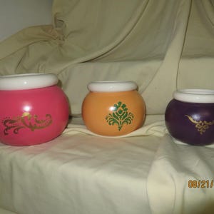May include: Three ceramic pots in different sizes and colours. The largest is pink with gold filigree, the middle is orange with a green design, and the smallest is purple with gold filigree. Each pot has a white rim.