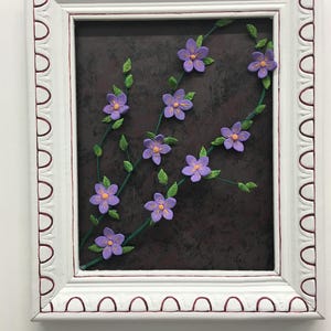 May include: A framed floral artwork featuring purple flowers with orange centers and green leaves on dark brown background. The white frame has a decorative edge with a red accent.