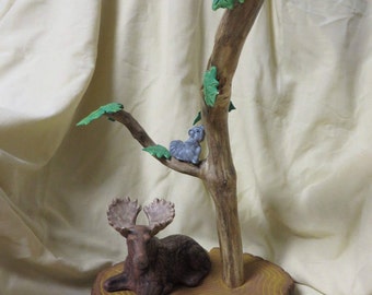May include: A ceramic moose figurine with large antlers is sitting on a brown tree stump base. A small grey squirrel figurine is climbing a brown tree branch with green leaves.