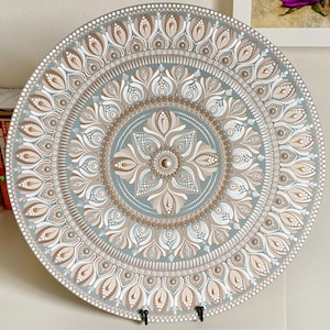 May include: A round, gray painted wooden plate with a white and brown mandala design. The mandala features intricate patterns of flowers and dots.