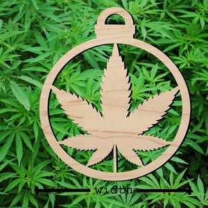 May include: A wooden ornament featuring a cannabis leaf design within a circular frame. The ornament is light brown and set against a background of green cannabis plants. The word "width" is written at the bottom.