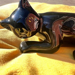 May include: A glossy black ceramic cat figurine with yellow and white accents. The cat is in a resting pose, with its head turned to the side. The figurine is on a yellow surface.