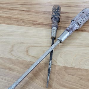 May include: Two vintage hand tools with weathered wooden handles and metal shafts. One tool features a flat-head screwdriver tip, while the other has a pointed, tapered metal tip. Both tools show signs of wear, including chipped paint and metal oxidation.