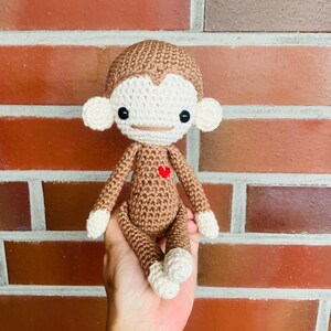 Könnte beinhalten: Eine handgefertigte Häkelpuppe in Braun- und Cremetönen. Der Affe hat ein herzförmiges Detail auf der Brust. Die Puppe hat ein lächelndes Gesicht, große Ohren und weiße Akzente an Händen und Füßen. Der Hintergrund ist eine Backsteinmauer.