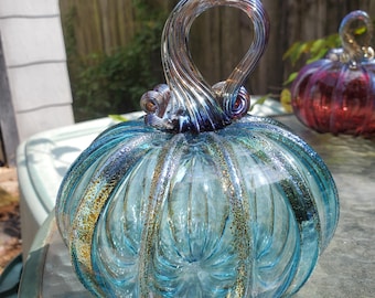May include: A hand-blown glass pumpkin in a vibrant light blue hue, featuring vertical ribbed detailing and a speckled gold-toned finish along the ridges. The piece is topped with a tall, curved, dark blue and metallic-toned glass stem.