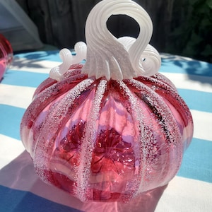 May include: A pink glass pumpkin with a white, swirled stem. The pumpkin has a textured surface with white speckles. The glass is transparent, allowing light to pass through. A decorative fall accent.