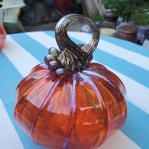 May include: A decorative glass pumpkin with an orange hue and iridescent highlights. The pumpkin features a detailed, swirled handle in a metallic finish. The pumpkin is sitting on a blue and white striped surface, suggesting a seasonal or autumnal theme.