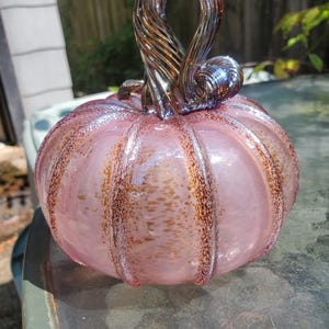 May include: A hand-blown glass pumpkin featuring a translucent light pink body with vertical ribs accented by speckled brown and gold flecks. The top includes a twisted, dark metallic glass stem that curls to one side, creating a decorative object.