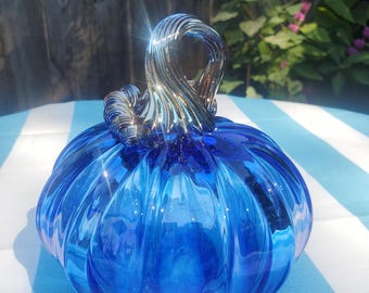 May include: A vibrant blue glass pumpkin with a swirled, silver-toned stem. The pumpkin has a ribbed design and sits on a blue and white striped surface. The glass reflects light, creating a shimmering effect.