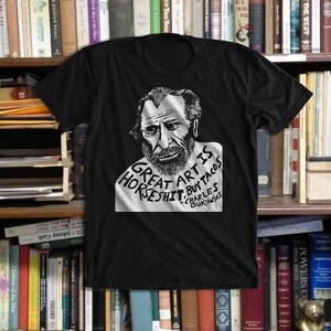 Puede incluir: Camiseta negra con un retrato en blanco y negro de Charles Bukowski y el texto "Great Art Is Horseshit, Buy Tacos" en estilo manuscrito. Cuello redondo.