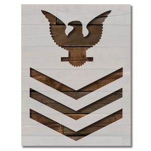 May include: A stencil of a US Navy Chief Petty Officer rank insignia. The stencil features a stylized eagle with wings spread and a chevron with three stripes. The design is cut out of a white material, revealing a wood grain background.