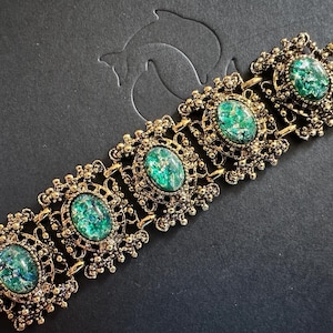 Puede incluir: Una pulsera vintage dorada con cinco cabujones ovalados verdes moteados. La pulsera presenta detalles de filigrana ornamentados alrededor de cada piedra y a lo largo de los eslabones de conexión. El cierre es de estilo simple de gancho y barra.