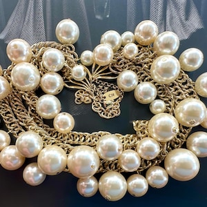 Puede incluir: Un collar de múltiples cadenas con grandes perlas falsas y eslabones de cadena dorados. El collar tiene una pequeña etiqueta con las letras "CH" en ella.