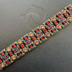 Puede incluir: Un brazalete vintage con base de metal dorado, con piedras preciosas rojas, naranjas y moradas. El brazalete tiene un diseño geométrico con piedras rectangulares y ovaladas. El cierre es visible en el lado izquierdo.