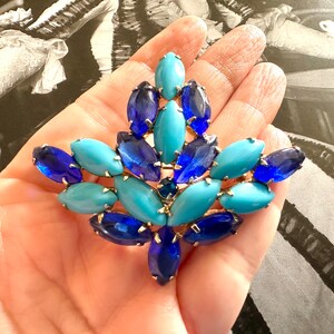 Puede incluir: Broche vintage con diseño floral y piedras preciosas azules y turquesas. El broche tiene un engaste de metal dorado. Las piedras son de forma ovalada y marquesa, dispuestas en un patrón simétrico.