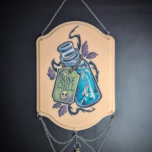 Può includere: Un'appendice murale decorativa che presenta una bottiglia di vetro blu con un osso all'interno, circondata da viti e foglie. La bottiglia ha un'etichetta che recita "BONE CHILL" con un grafico di teschio. L'appendice è attaccata a una catena con tre gocce di vetro blu e un piccolo flacone di vetro.