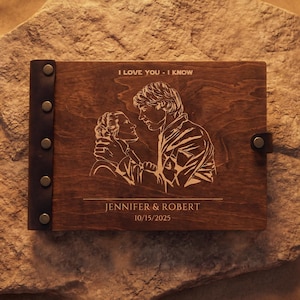 Könnte beinhalten: Ein rechteckiges Fotoalbum aus Holz mit dunkelbrauner Oberfläche, das eine Lasergravur eines Paares, den Text I LOVE YOU - I KNOW, JENNIFER & ROBERT sowie 15.10.2025 zeigt. Das Album hat einen dunklen Lederrücken, der mit fünf Metallnieten befestigt ist.