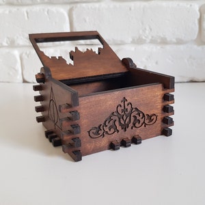 Pu&ograve; includere: Una scatola di legno con un coperchio incernierato, che presenta un design intagliato di un castello sul coperchio e un motivo floreale decorativo sul fondo. La scatola &egrave; di colore marrone e ha una finitura in legno scuro. Il lato della scatola presenta un'immagine intagliata del simbolo dei Doni della Morte.